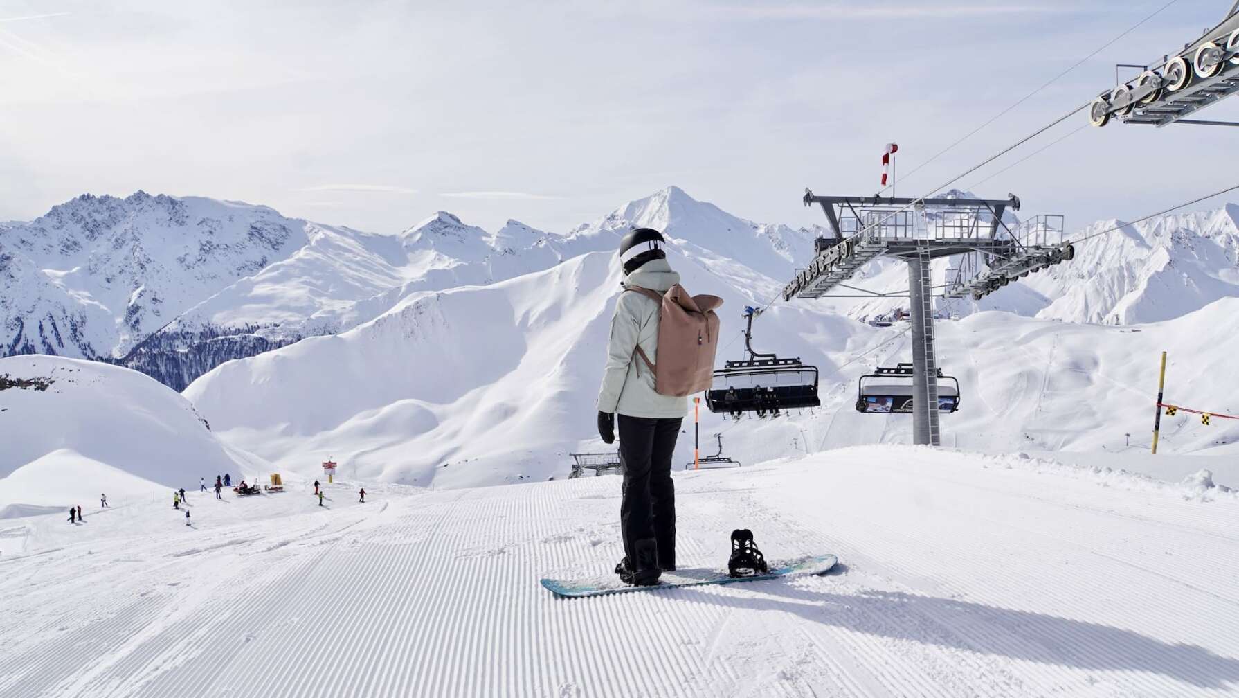 Foto eines Snowboarders vor dem herrlichen Panorama der schneebedeckten Berge in der Silvretta Arena in Samnaun/Ischgl, am Bildrand ein Sessellift